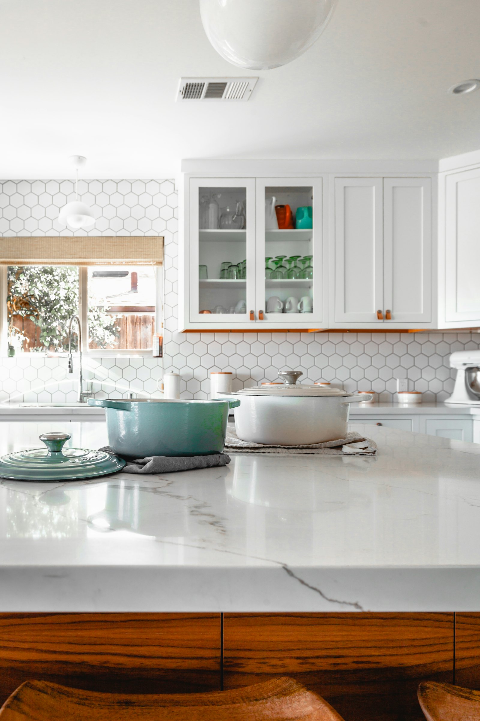 A sunlit kitchen with white cabinets, a marble island, and cookware on the counter.